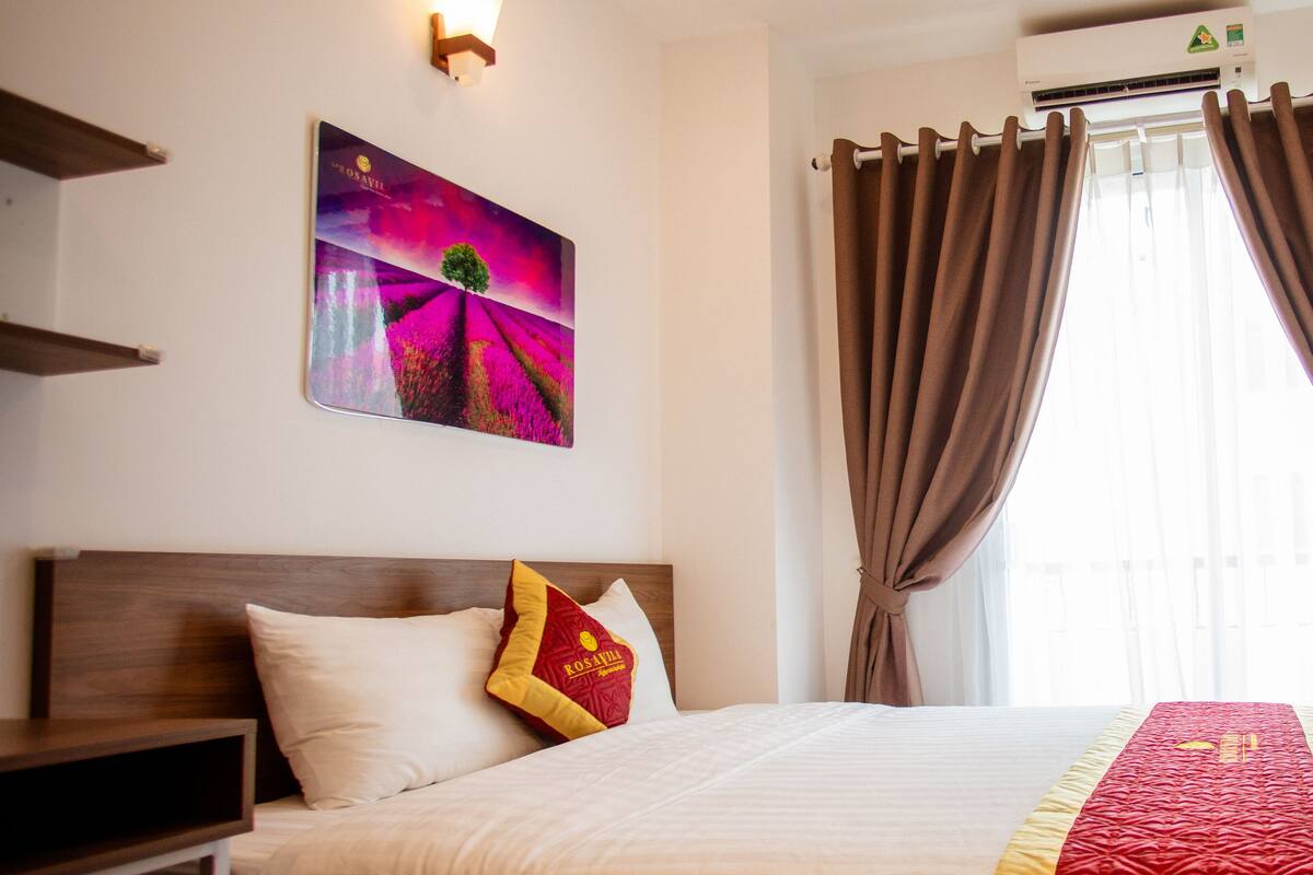 A hotel room with a bed, a nightstand, a wall-mounted painting, a window with curtains, and an air conditioning unit.