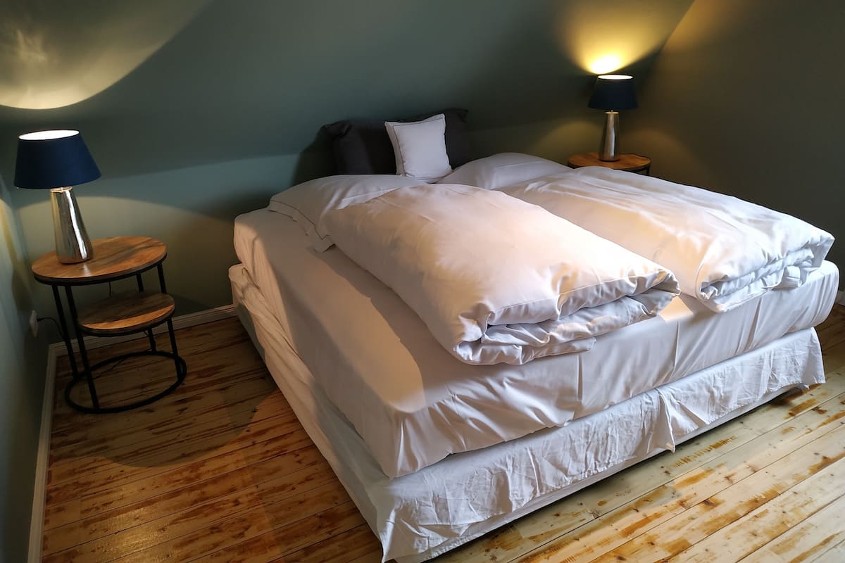 Ein ordentlich bezogenes Bett mit weißen Leinentüchern, zwei Nachttischlampen und ein kleiner Holztisch daneben.