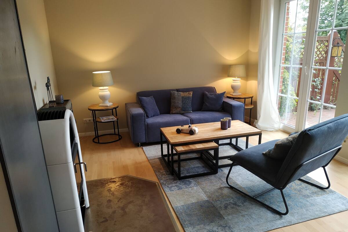 Ein modernes Wohnzimmer mit einer Couch, einem Couchtisch und einem Sessel. Zwei Lampen stehen auf Beistelltischen, und ein Fenster bietet Blick auf grüne Vegetation draußen.