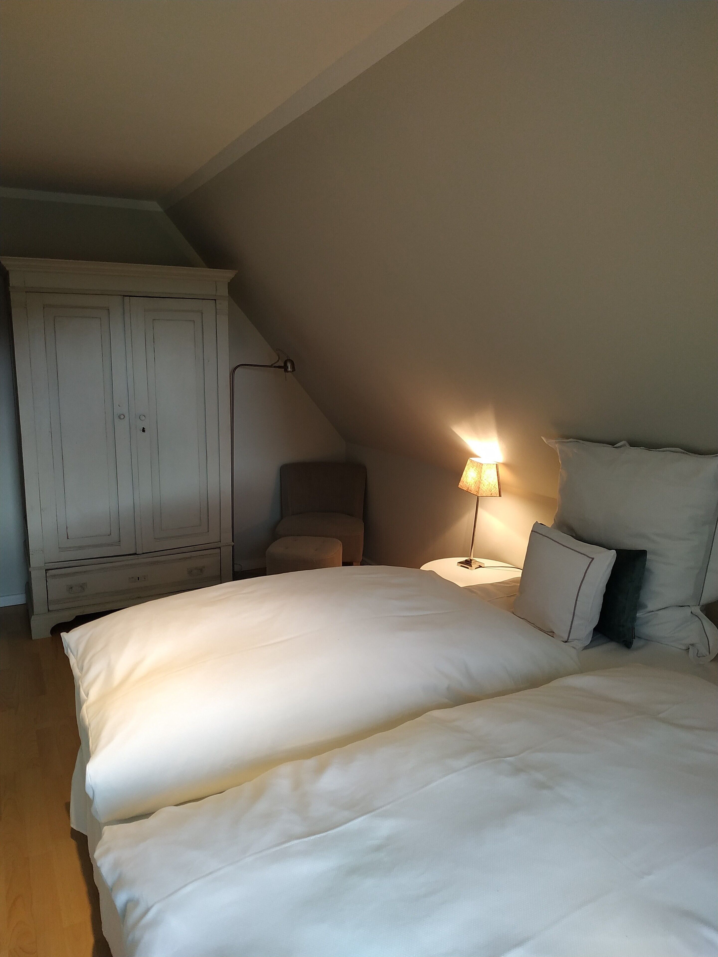 A bedroom with a large bed, a chair, a wardrobe, and a nightstand with a lamp.