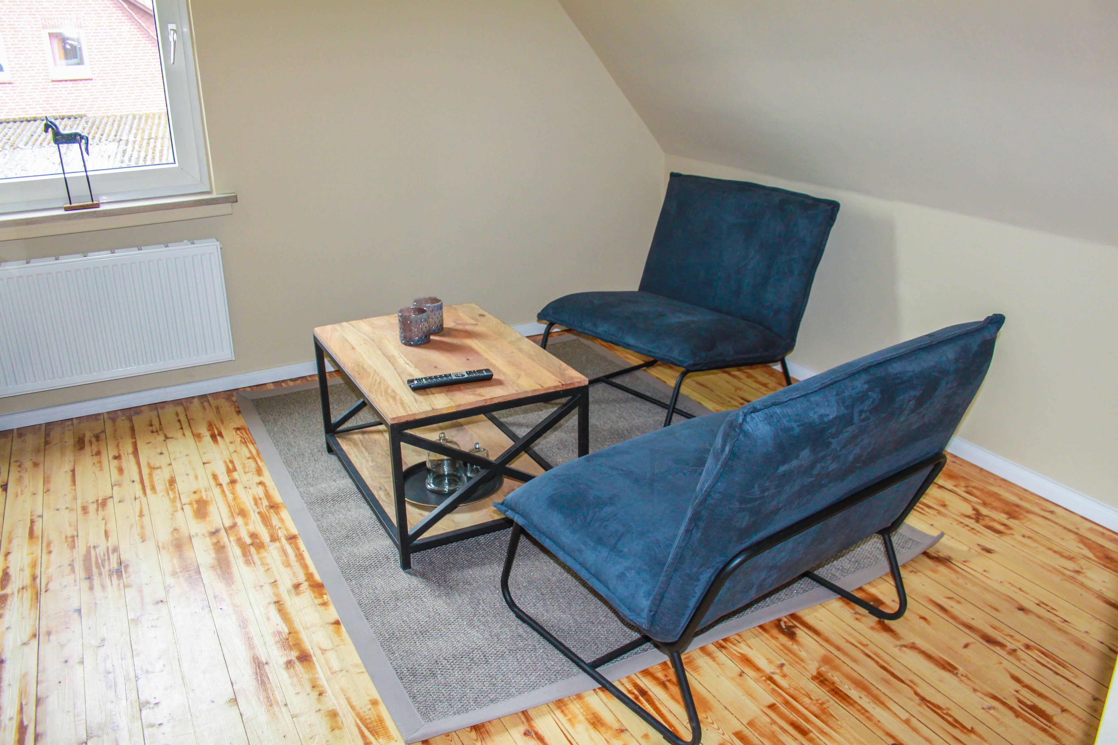 A small room with a sloped ceiling, a wooden floor, a small table with a remote control, and two blue chairs.