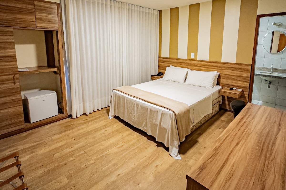 A hotel room with a bed, bedside table, lamp, wooden floor, and a mirror.
