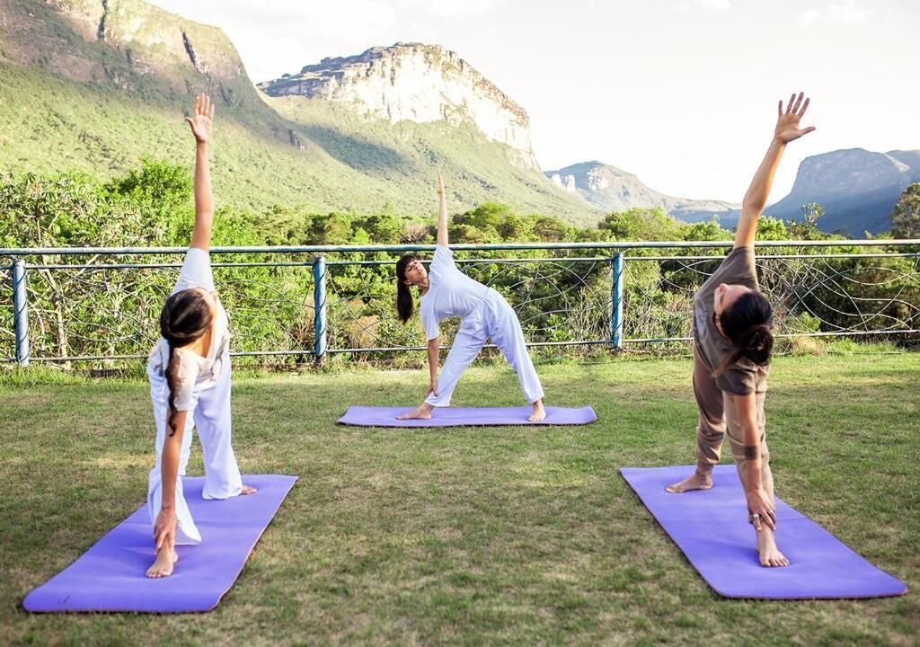 Drei Personen üben Yoga auf violetten Matten im Freien, mit Bergen im Hintergrund.