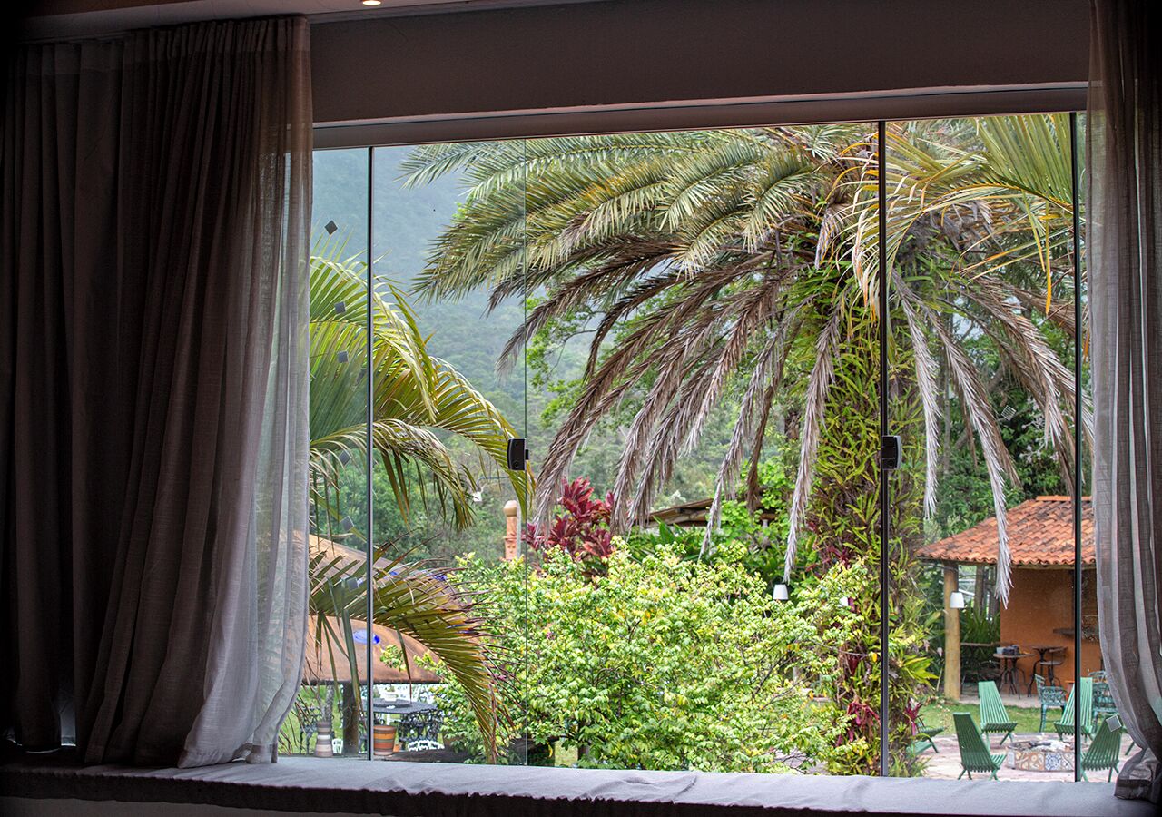 Una habitación con un gran ventanal que da a un jardín tropical con palmeras y una estructura de techo de paja.