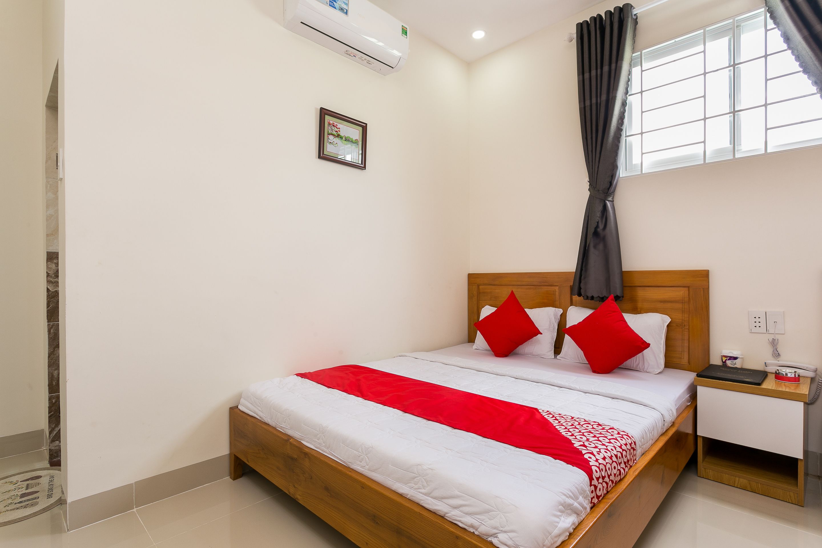 A hotel room with a wooden bed, white bedding, a red throw, a nightstand with a phone, and a window with curtains.