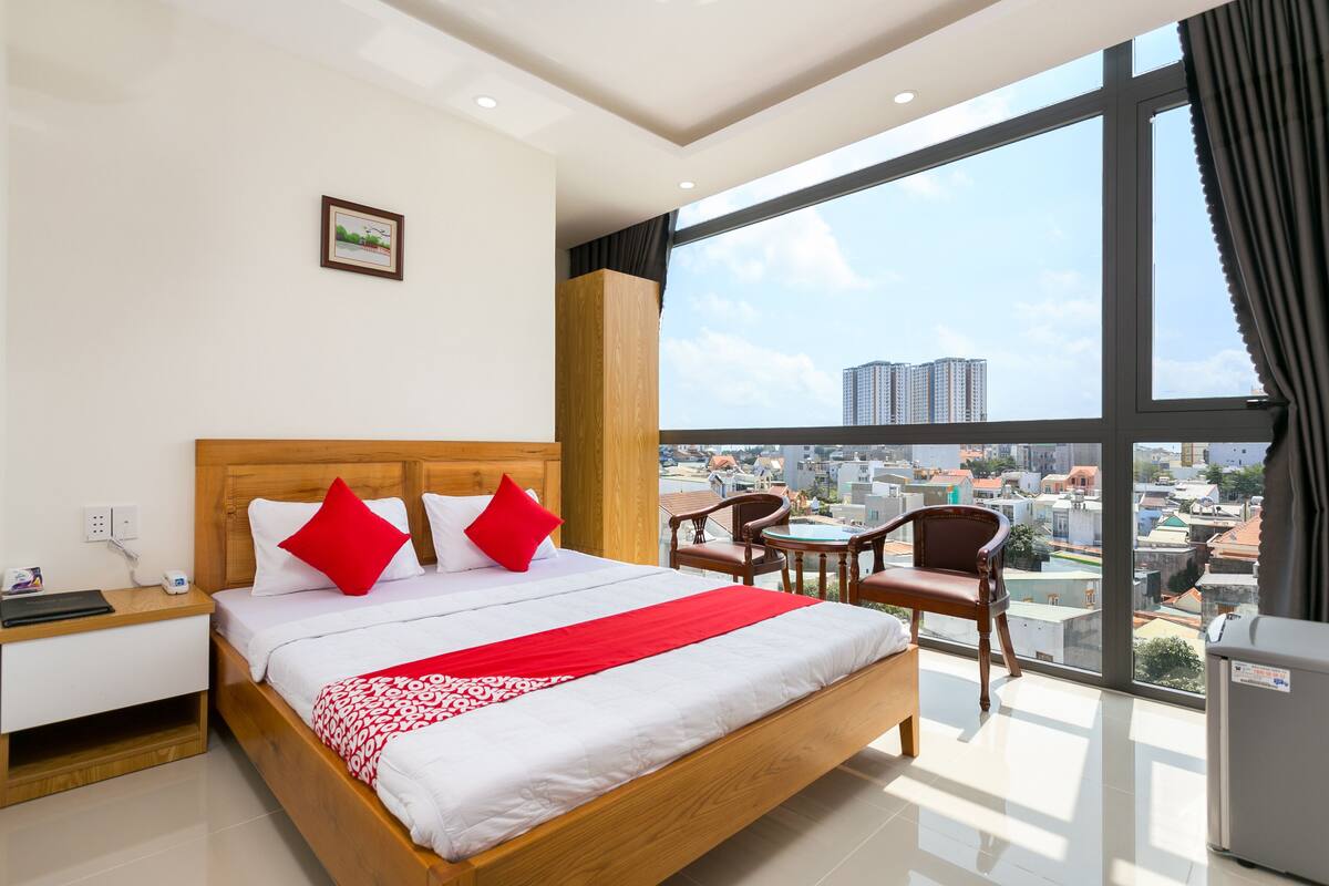 Habitación de hotel con una cama grande, un escritorio, una silla y vistas de la ciudad a través de amplias ventanas.