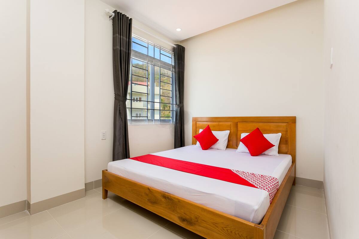 Una cama bien hecha con tablero de madera, ropa de cama blanca y detalles en rojo, ubicada en una habitación con un gran ventanal y cortinas.