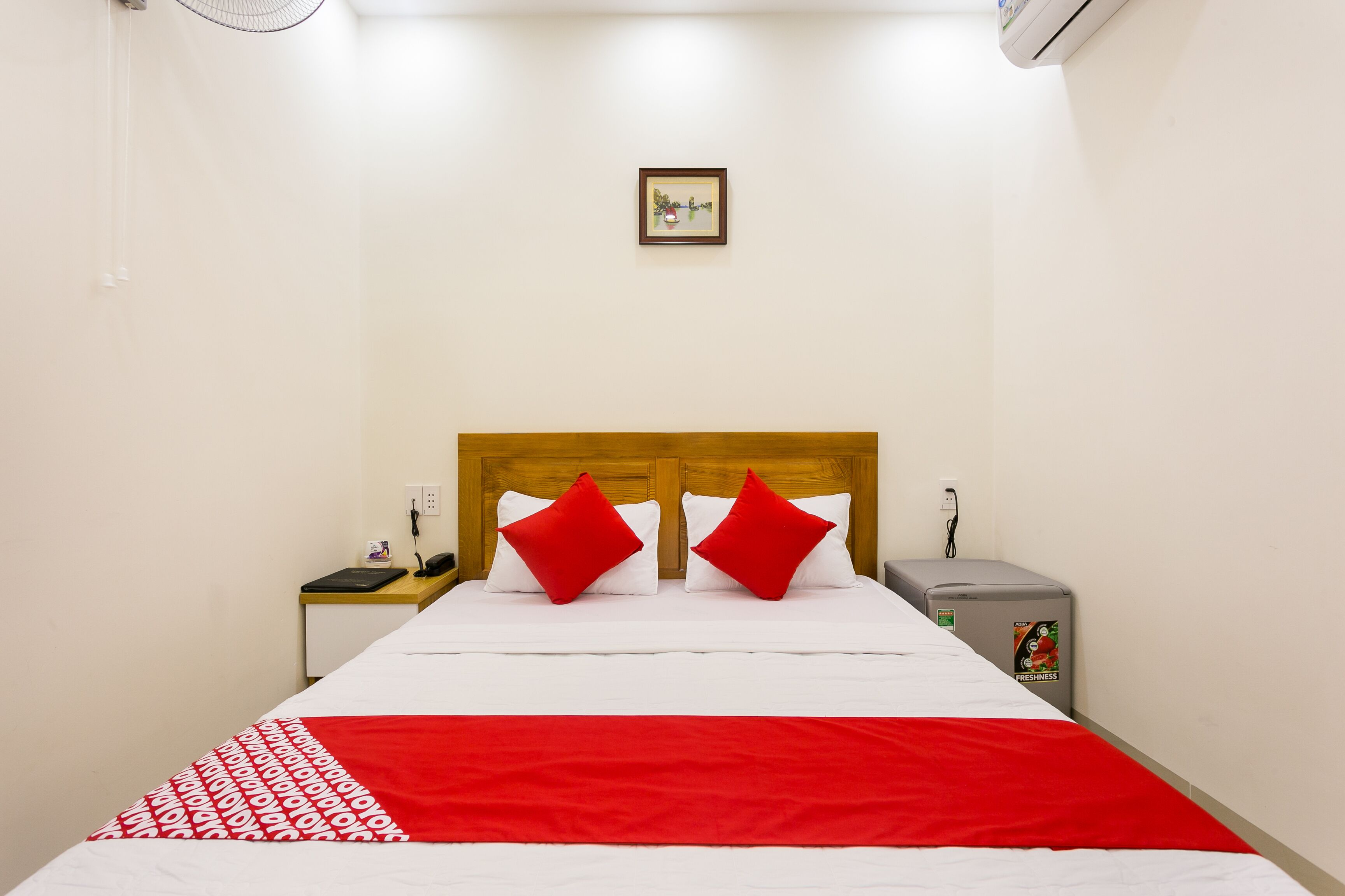Una cama bien hecha con colcha blanca y roja, dos almohadas rojas y un cabecero de madera. A la derecha de la cama, hay una mesita de noche con una lámpara, un enchufe y un refrigerador pequeño.