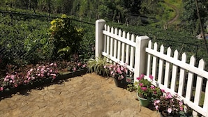 A paved area with a white picket fence, surrounded by lush greenery and flowering plants. - Windsor Forest Bungalow (Bandarawela)