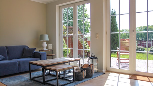 A living room with a sofa, a coffee table, a lamp, and a view through a glass door to an outdoor area with a wooden deck and greenery. - Ferienapartments Lüneburger Heide - St. Dionys (Barum)