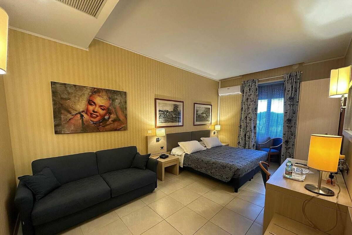Una habitación de hotel con cama, sofá, escritorio y dos lámparas. Se observan cuadros en la pared y una ventana con cortinas.