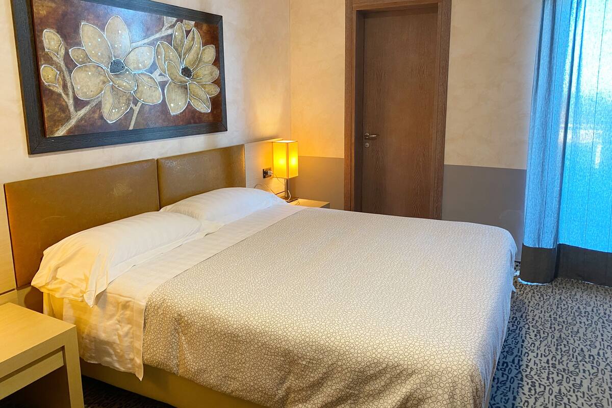 Habitación de hotel con cama, mesita de noche, lámpara, puerta y un cuadro en la pared.