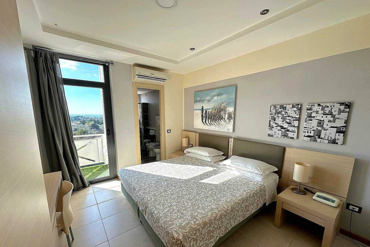Habitación de hotel con una cama grande, una mesita de noche con lámpara, vista a la ciudad por la ventana y obras de arte en la pared.