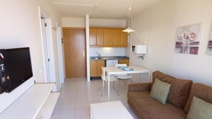 Un salon avec un canapé marron, une table basse blanche, un téléviseur et un coin cuisine doté de meubles en bois et d’un plan de travail blanc.
