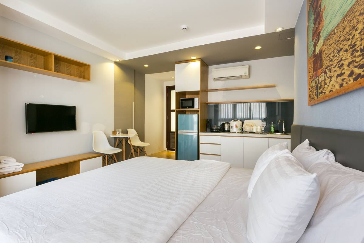 A hotel room with a bed, a desk, a chair, a television, a kitchenette, and a painting on the wall.