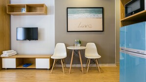 Een moderne hotelkamer met een houten bureau, een flatscreen-tv, een kleintje tafeltje met twee witte stoelen, een ingelijst foto van een strand en een blauwe koelkast.