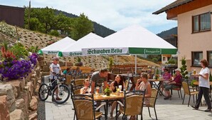 Un café en plein air avec des tables et des chaises, une tente blanche arborant le logo « Schwaben Brau », et un bâtiment agrémenté de jardinières fleuries.