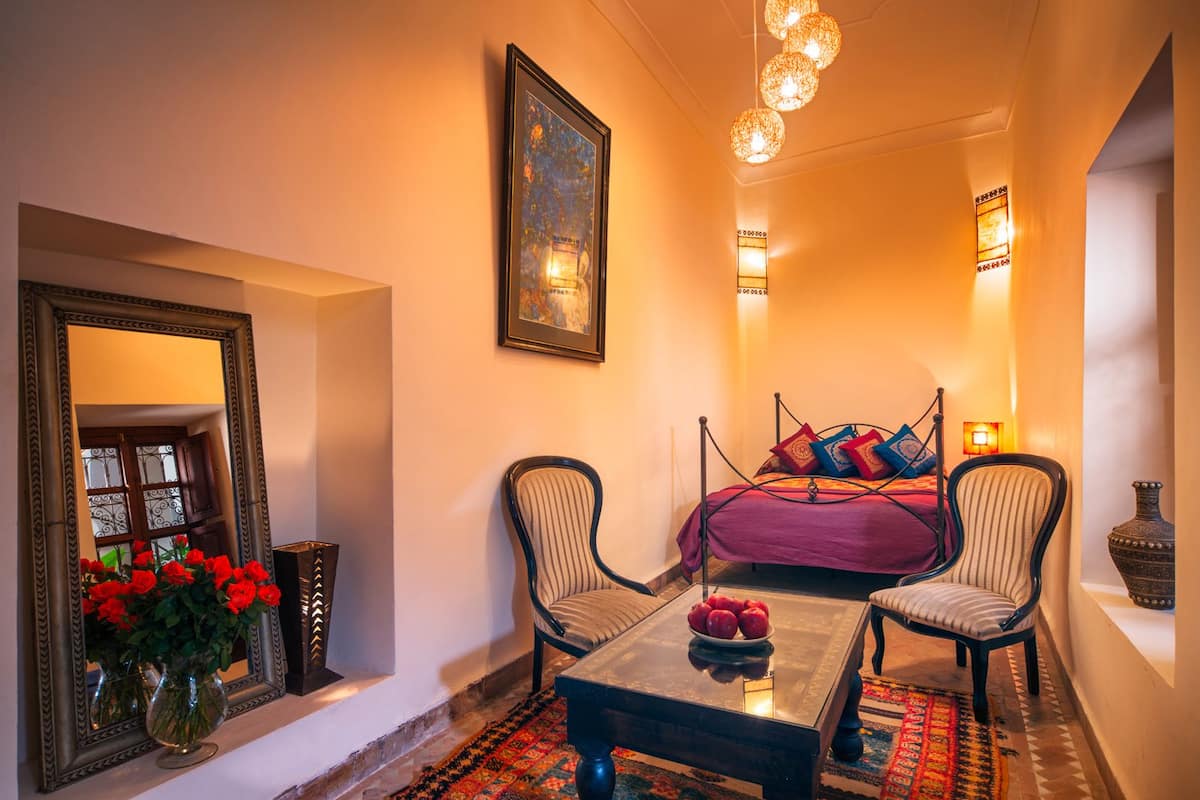 A cozy room with a bed, two chairs, a table, a mirror, and a vase of red flowers.