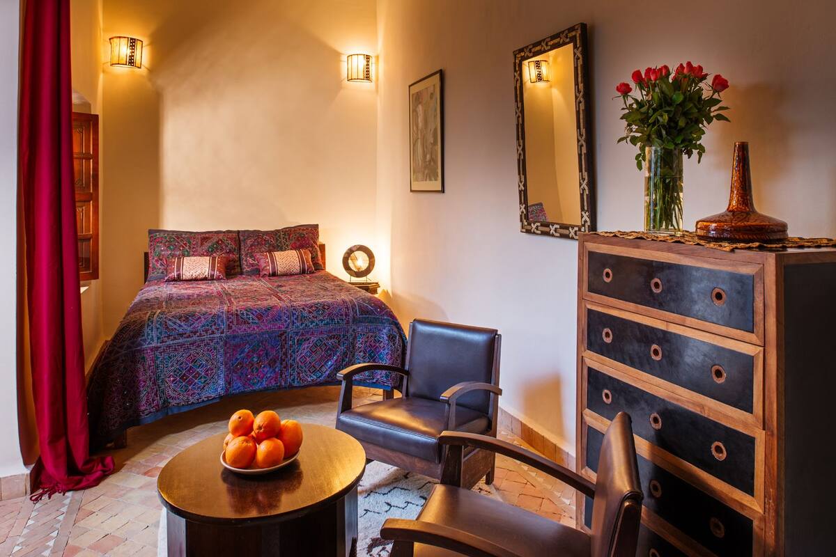 A bedroom with a bed, two chairs, a dresser, a mirror, and a vase with flowers.