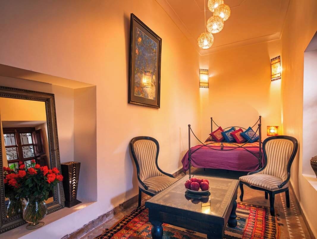 A hotel room with a bed, two chairs, a table with fruit, a mirror, and decorative lights.