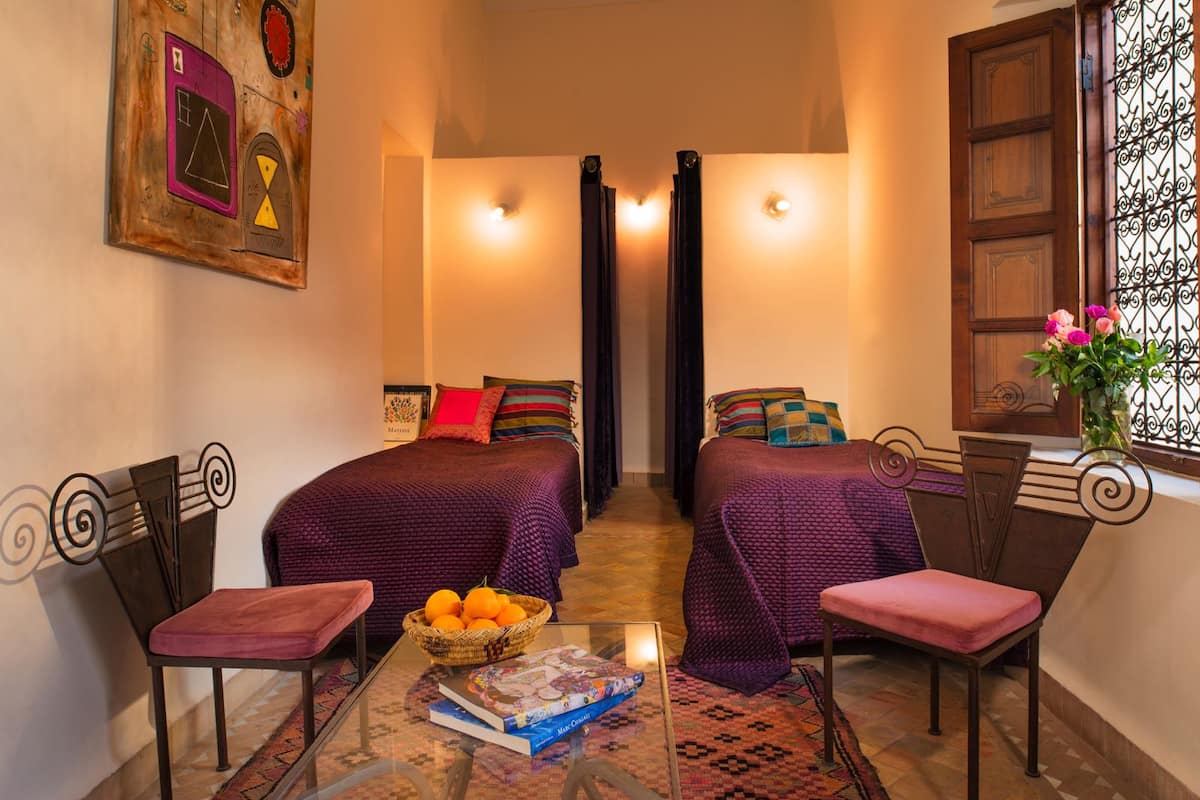 A cozy room with two beds, a small table with a book and oranges, and a window with flowers.