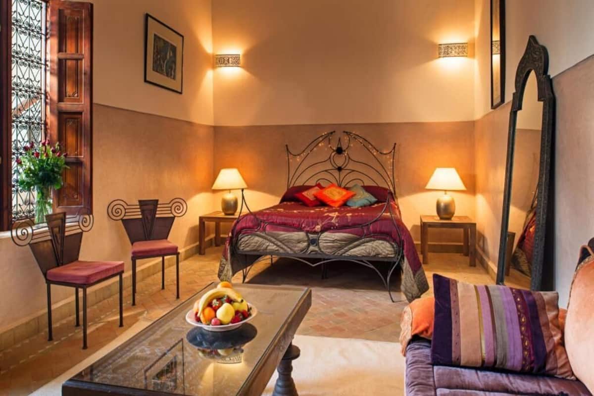A bedroom with a bed, two bedside tables with lamps, a coffee table with fruit, a sofa, and a mirror.