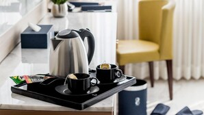 Habitación de hotel con televisión montada en la pared, una zona de café con hervidor, tazas y una bandeja, una silla amarilla y una ventana con cortinas.