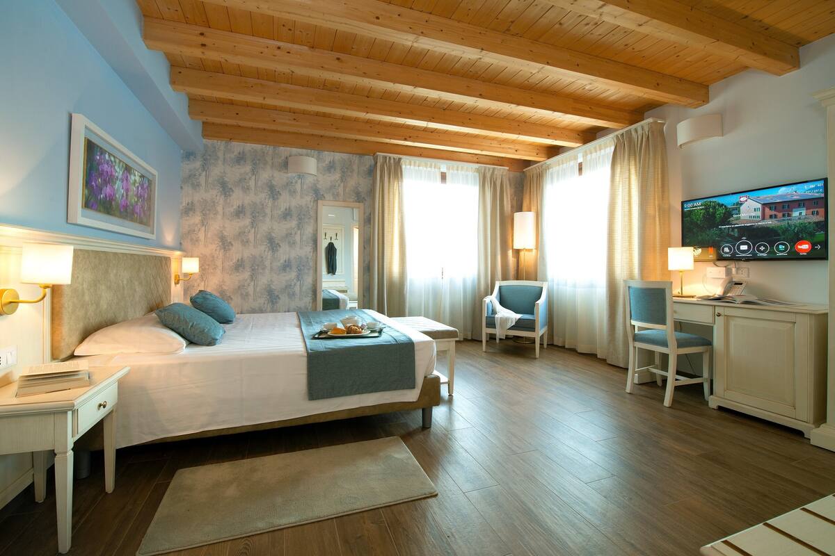 A hotel room with a wooden ceiling, a bed with pillows, a desk with a chair, a TV, and a window with curtains.