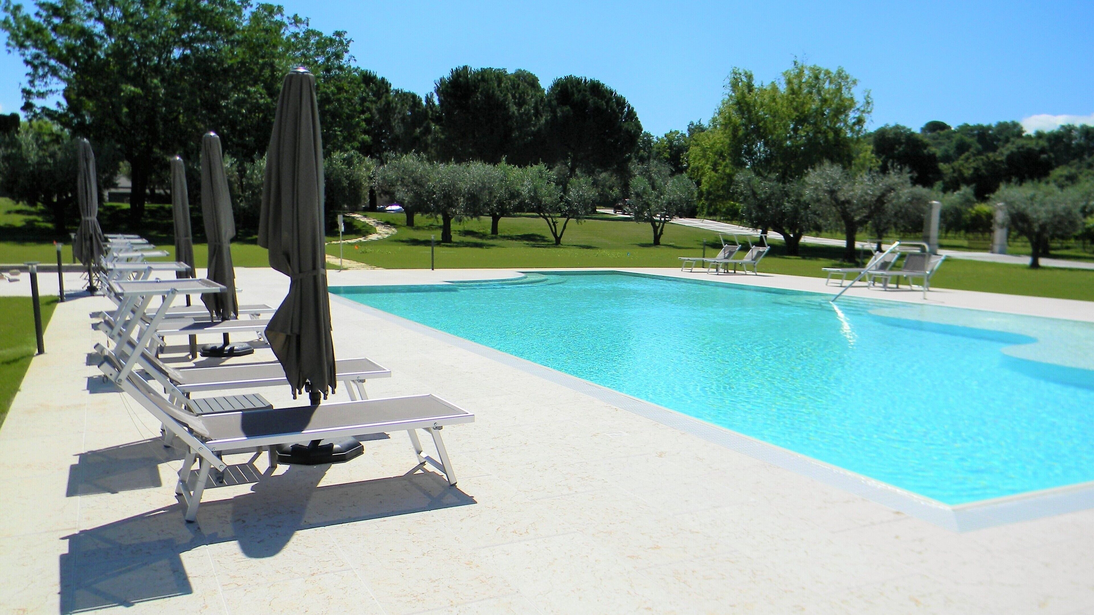A swimming pool with sun loungers and umbrellas, surrounded by trees and a grassy area.