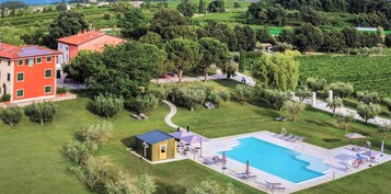 Una grande piscina circondata da lettini e ombrelloni, con un edificio rosso e una vegetazione rigogliosa sullo sfondo.