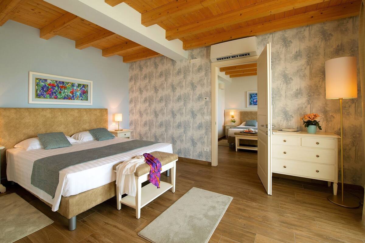 A bedroom with a large bed, bedside tables, a nightstand, a dresser, a floor lamp, and a painting on the wall.