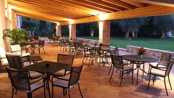 Un patio exterior con varias mesas y sillas, rodeado de pilares de ladrillo y vigas de madera.