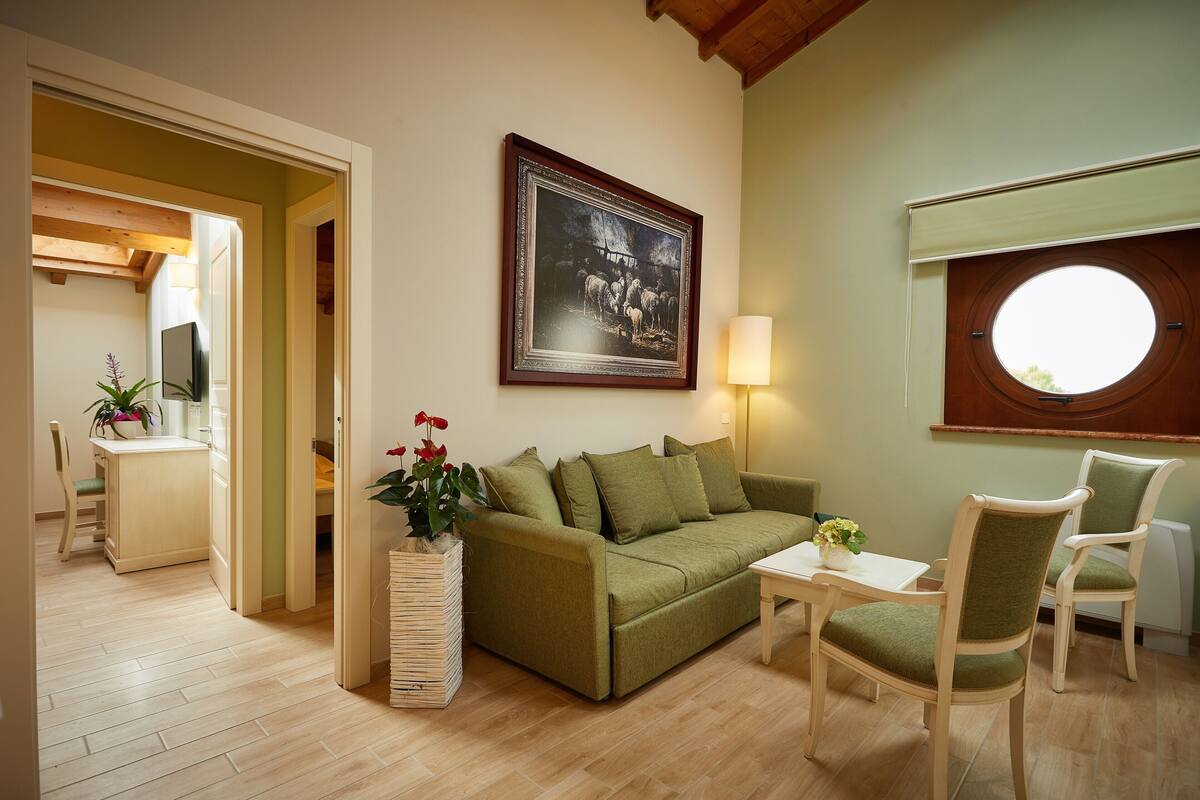 A cozy living room with a green sofa, two chairs, a small table, a potted plant, a framed picture on the wall, and a window with a view.