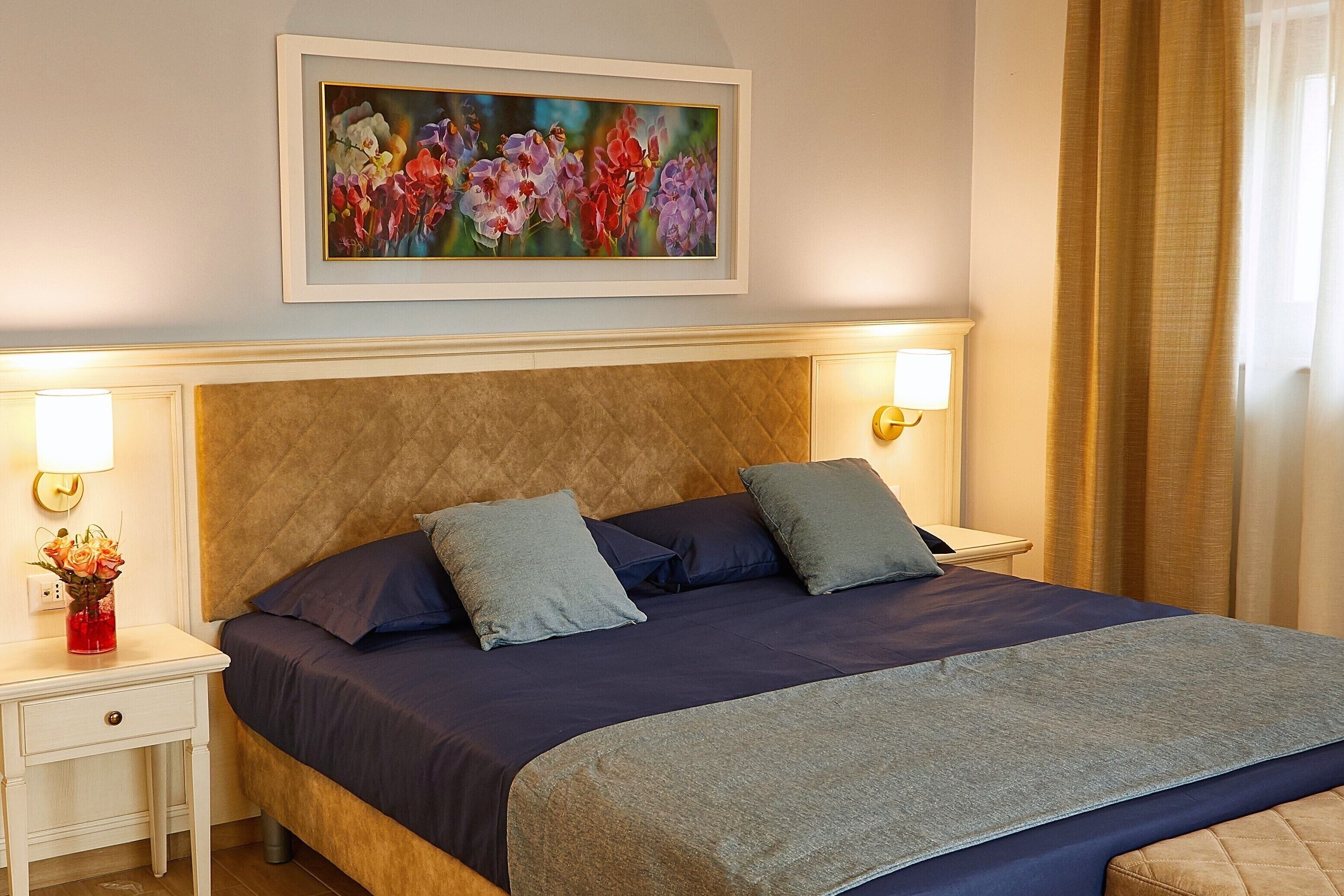 A neatly made bed with a quilted headboard, two bedside tables with lamps, and a framed floral artwork above the bed.