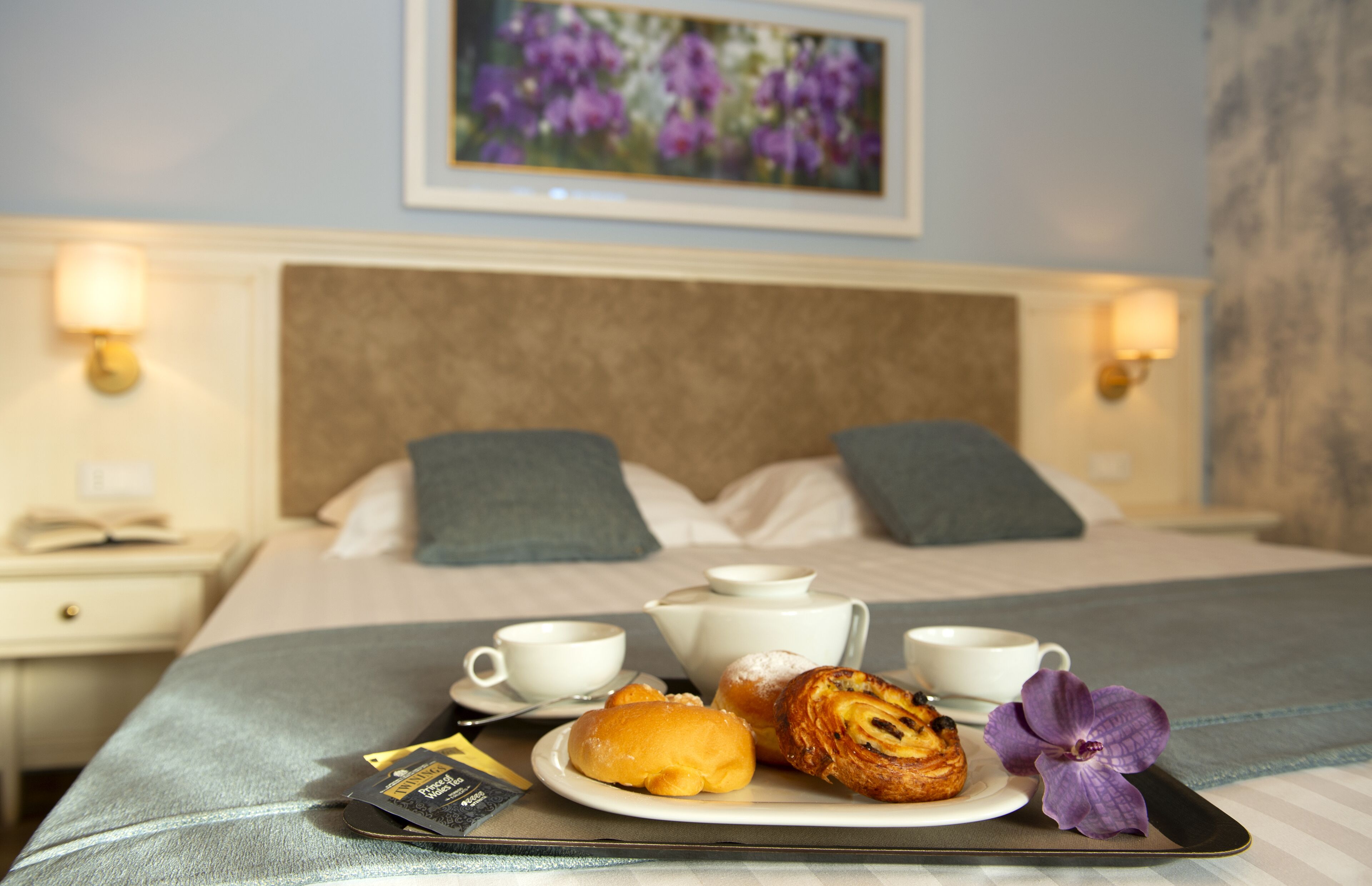 Una cama bien hecha con almohadas, una mesita de noche con lámpara y una bandeja de desayuno con pasteles y una flor.