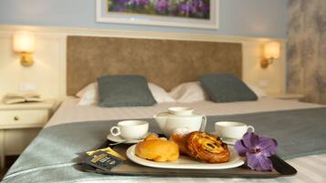 Una cama bien hecha con almohadas, una mesita de noche con lámpara y una bandeja de desayuno con pasteles y una flor.