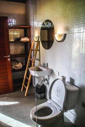 A bathroom with a toilet, sink, and a mirror. There is a wooden ladder leaning against the wall and a shelf with towels. - Kubu Jamu Ubud (Ubud)