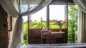 A bedroom with a bed, a wooden desk, a chair, a window with curtains, and a view of greenery outside. - Kubu Jamu Ubud (Ubud)