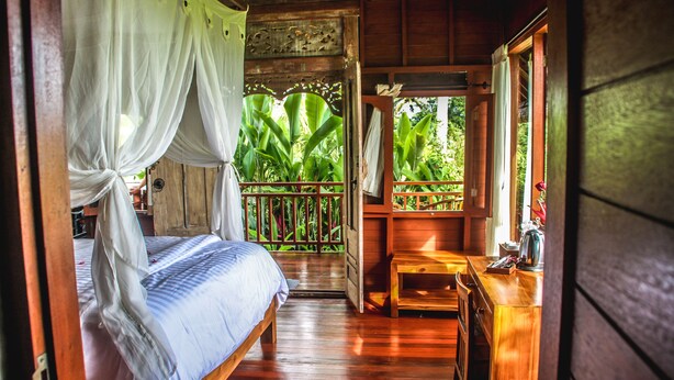 A wooden room with a bed, a wooden bench, and a view of lush greenery outside. - Kubu Jamu Ubud (Ubud)