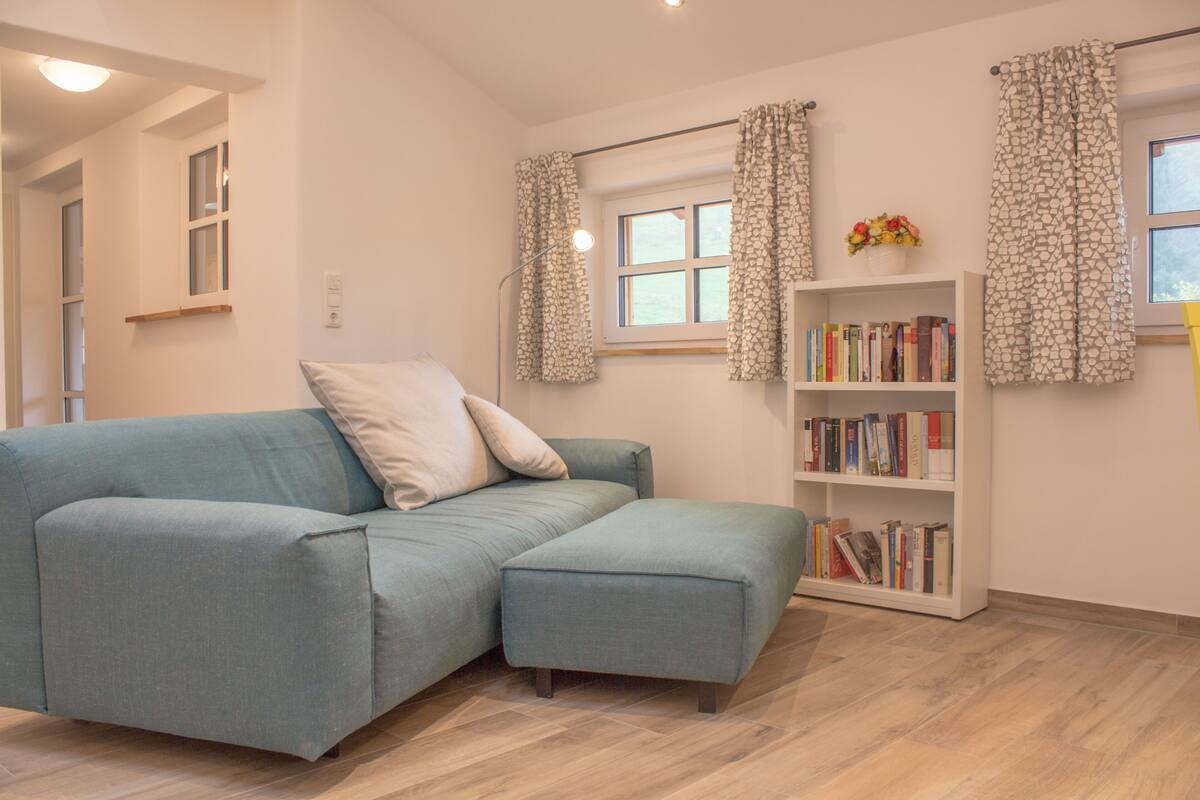 A modern living room with a sectional sofa, a bookshelf, and a window with patterned curtains.