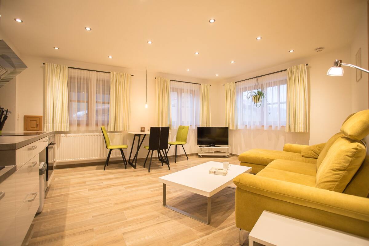 A modern living room with a yellow sofa, a white coffee table, and a TV. There is a dining area with a table and chairs, and a kitchen area with cabinets and a sink.