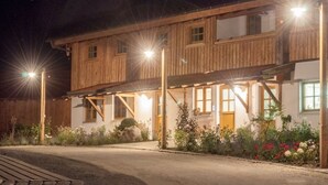 A two-story wooden building with a balcony, illuminated by exterior lights, surrounded by a garden with various plants and flowers. - Dauscher Hof (Siegsdorf)