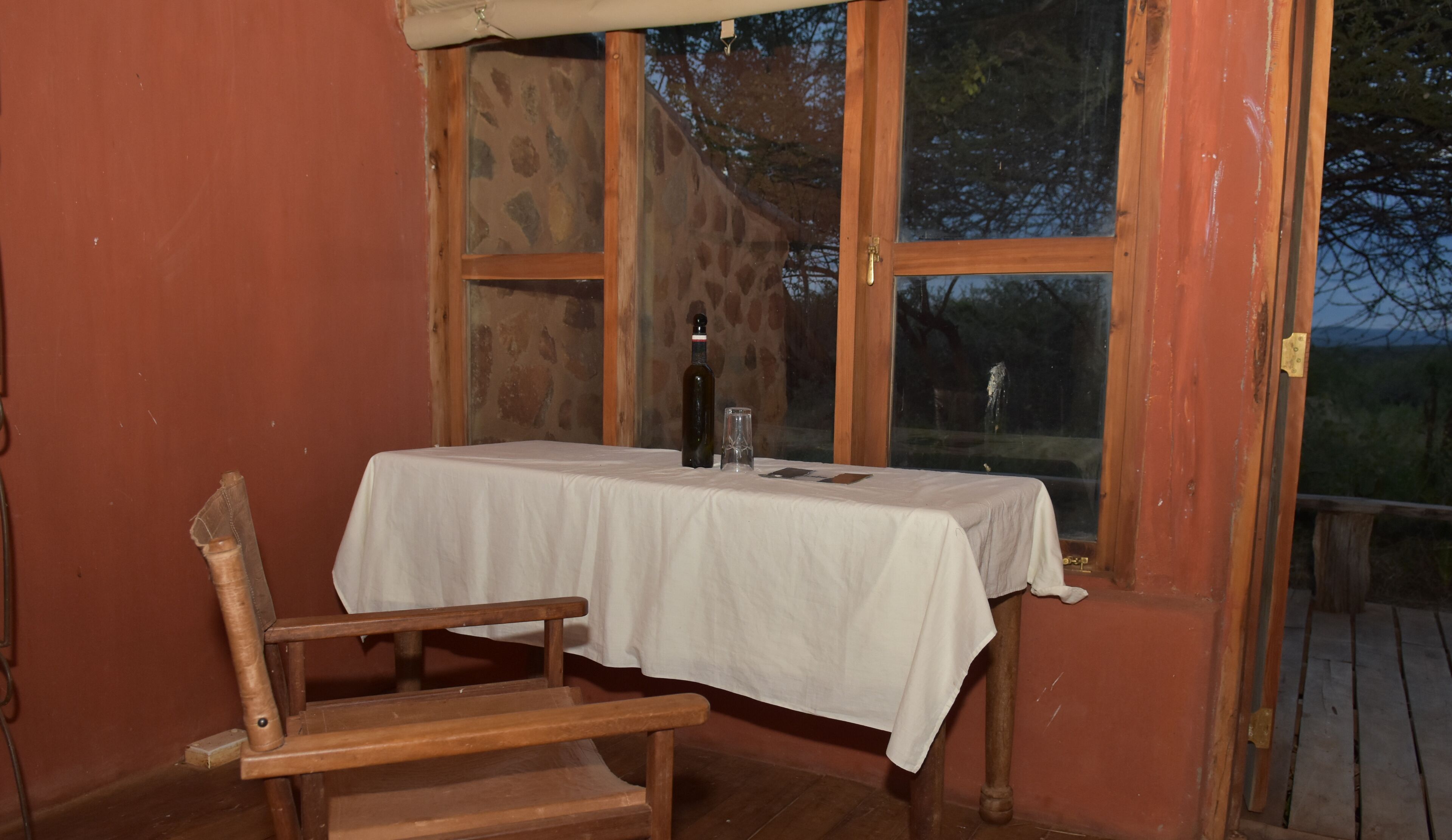 A small table with a white tablecloth, a bottle, and a glass on it, positioned near a window with a view of trees.