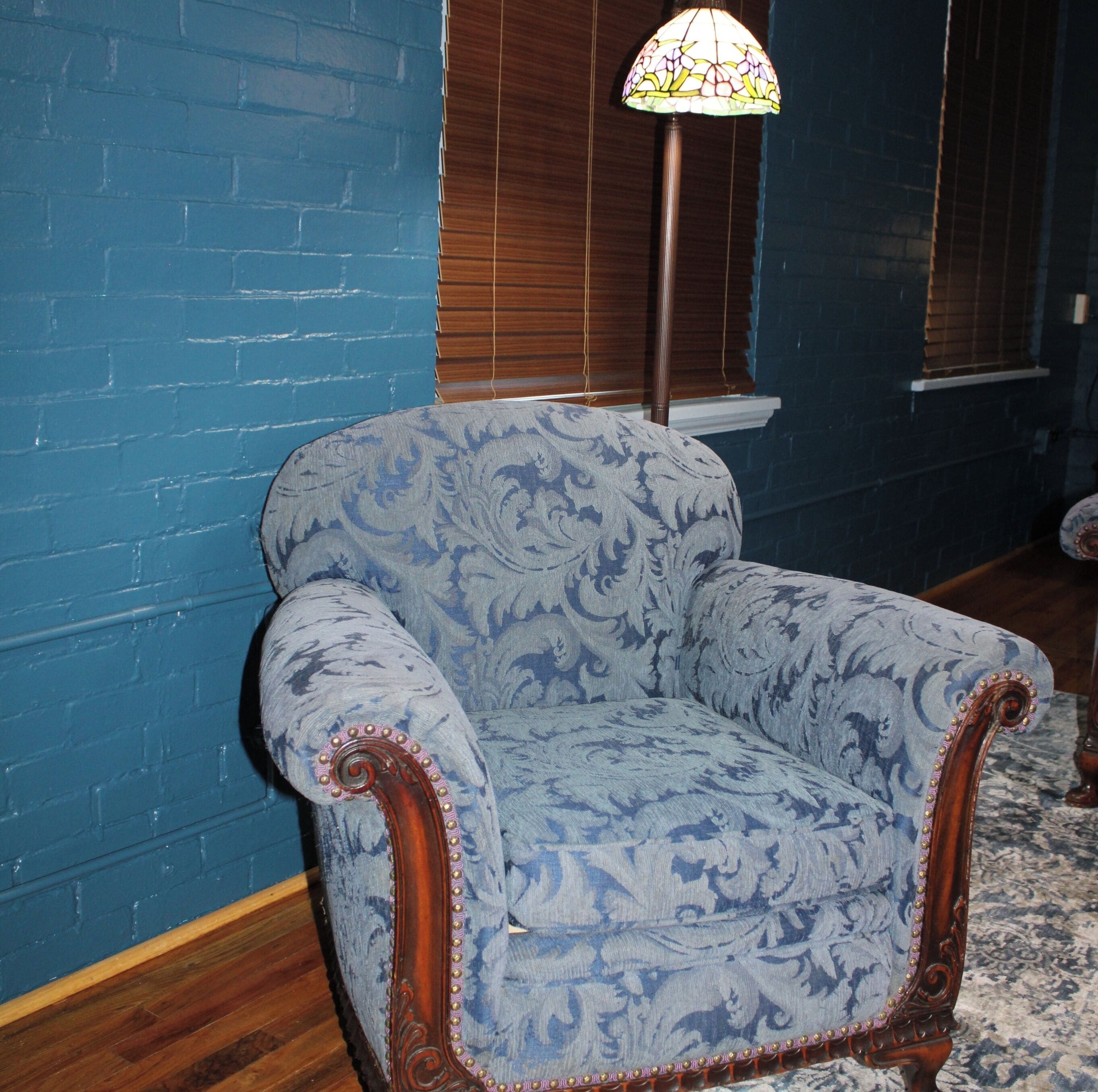 A blue patterned armchair with ornate wooden armrests and a matching floor lamp in a room with blue brick walls and wooden flooring.