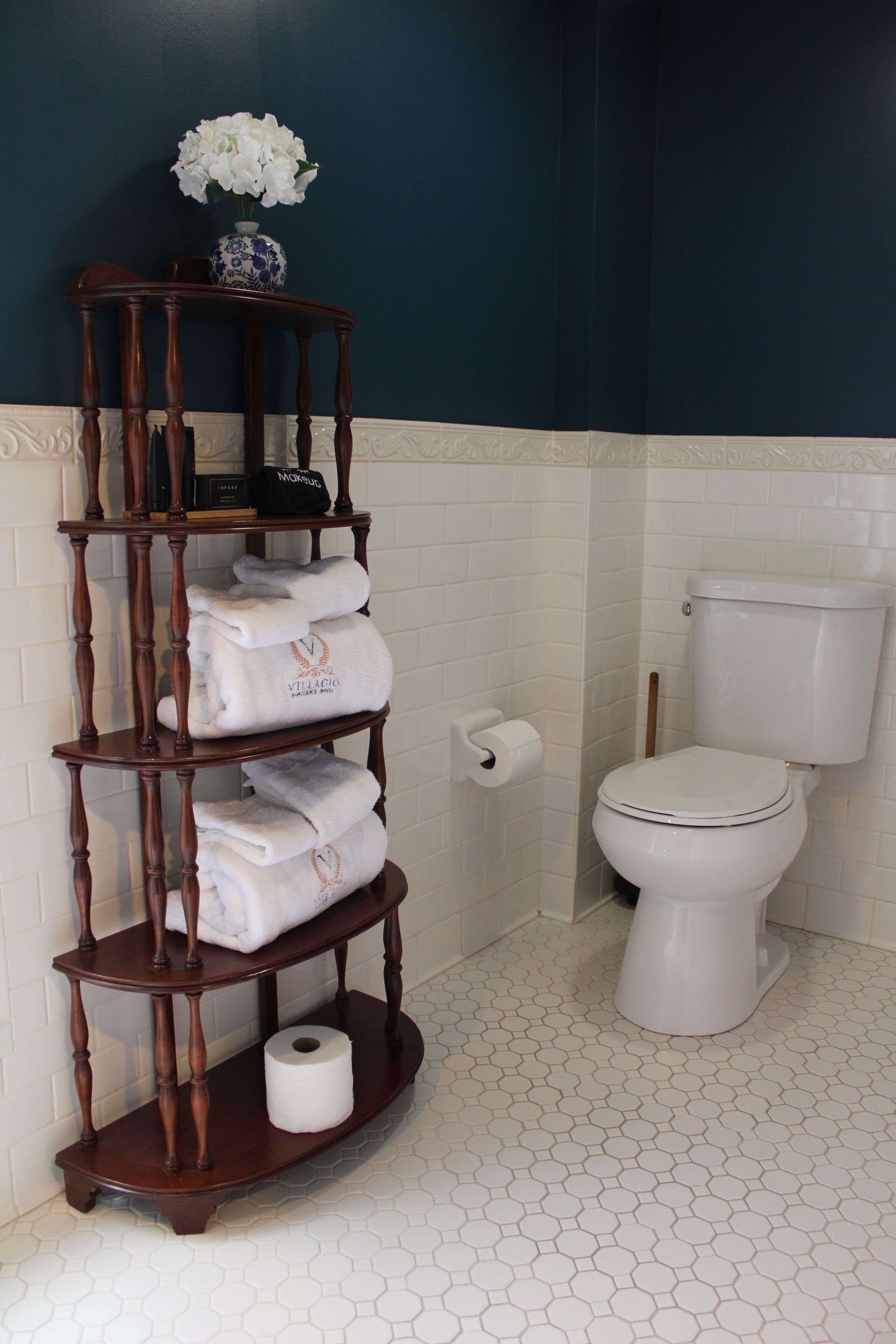 Une salle de bain avec des toilettes blanches, un porte-serviettes en bois sur lequel sont pliées des serviettes, un rouleau de papier toilette et un vase avec des fleurs blanches accroché au mur.