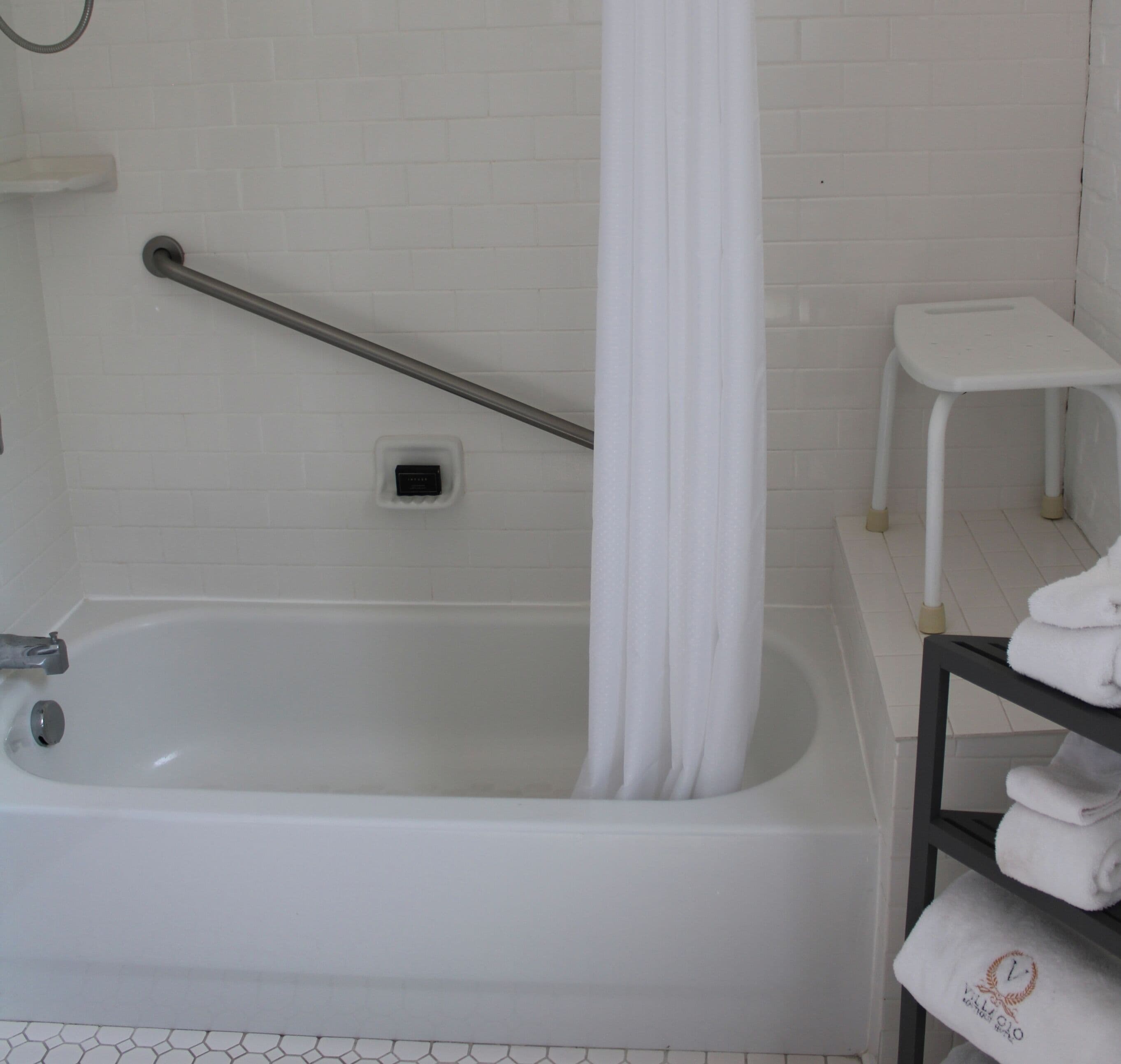 A white bathtub with a shower curtain, a grab bar, a small stool, and a towel rack with towels.