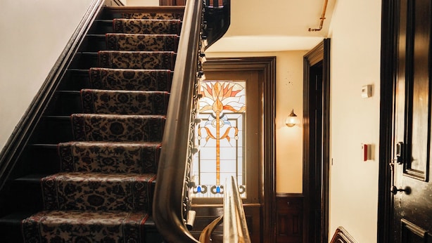 Una escalera con alfombra estampada, barandilla metálica y suelo de madera que conduce a una puerta con un vitral.