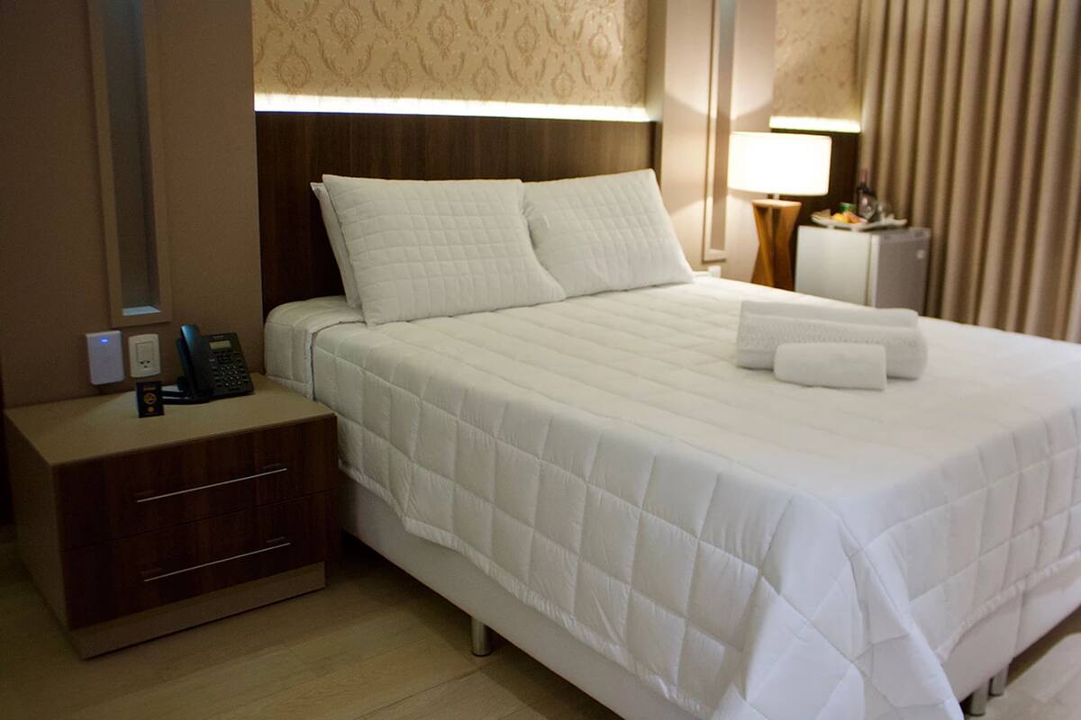 Quarto de hotel com uma cama bem arrumada, uma mesa de cabeceira com abajur e um pequeno armário.