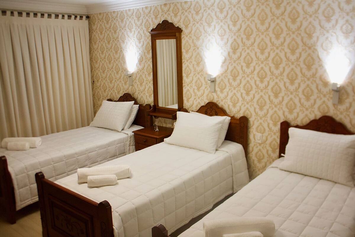 Quarto de hotel com duas camas de solteiro, cabeceira de madeira, mesa de cabeceira, espelho e arandelas.