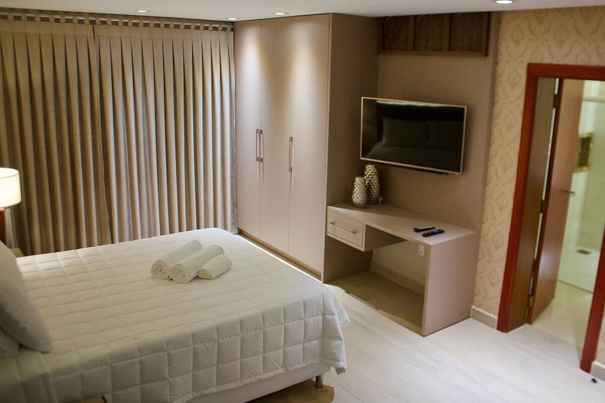 Quarto de hotel com cama, escrivaninha, televisão e guarda-roupa.
